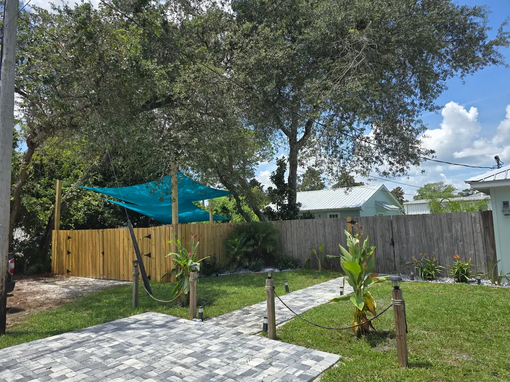 A bright backyard featuring a paved path, green lawn, wooden fence, blue shade sails, and trees, with houses in the background.