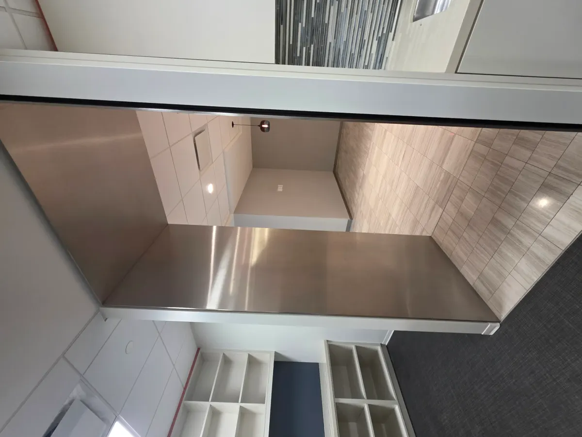Modern office interior with a long, reflective stainless steel counter, patterned tile, dark carpet, and white shelving units.