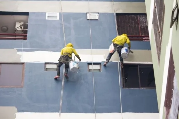 Two painters suspended by ropes are painting the exterior wall of a building dark blue and white, wearing yellow jackets.