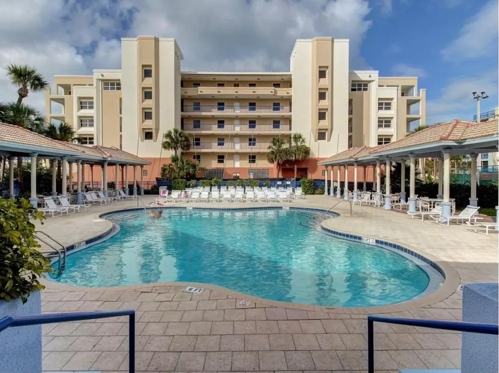 A resort complex featuring a large, winding swimming pool, numerous lounge chairs, shaded pergolas, and a multi-story building.