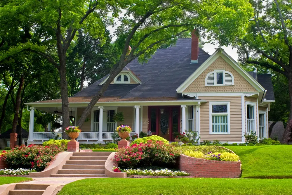 A large, classic home with a front porch, dark roof, and gabled windows, surrounded by lush trees and vibrant flower beds.