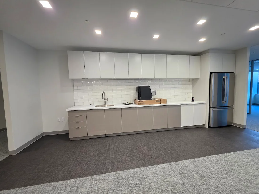 Sleek office kitchenette with white upper and light brown lower cabinets, subway tile, sink, Keurig, and stainless steel fridge.