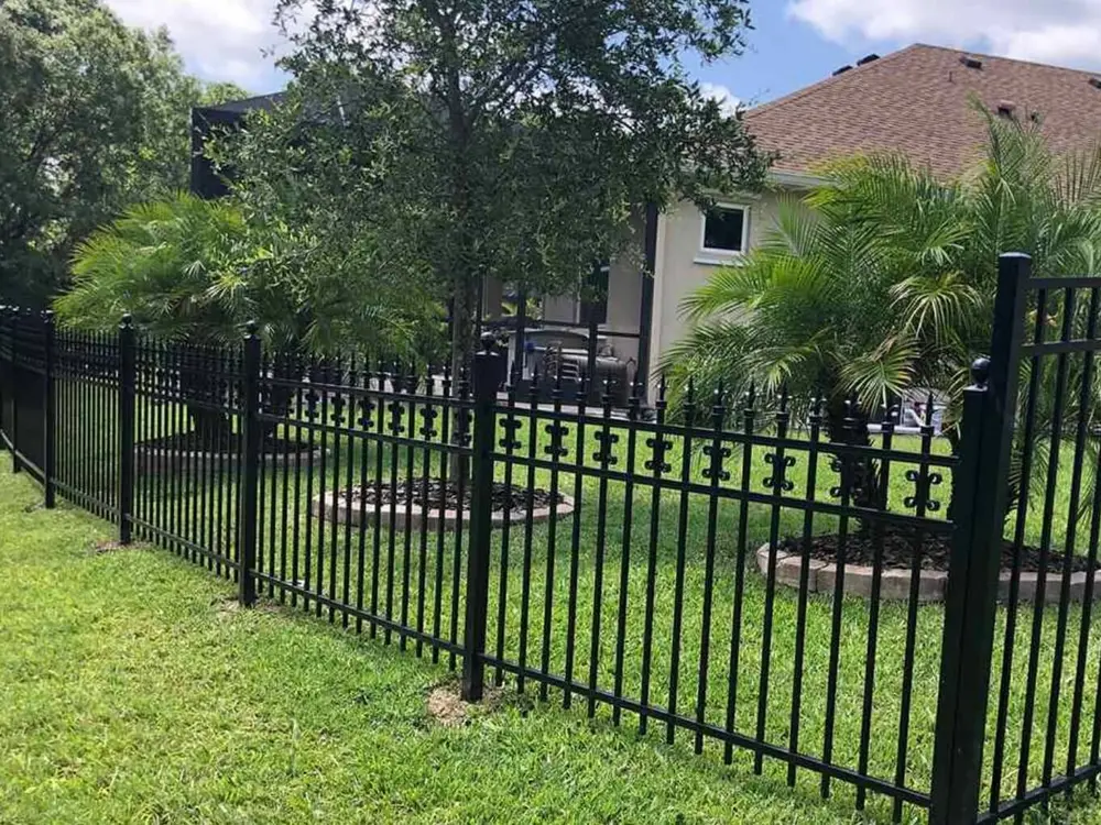 A modern black metal fence extends across a bright green lawn, featuring residential buildings and lush trees under a clear blue sky.