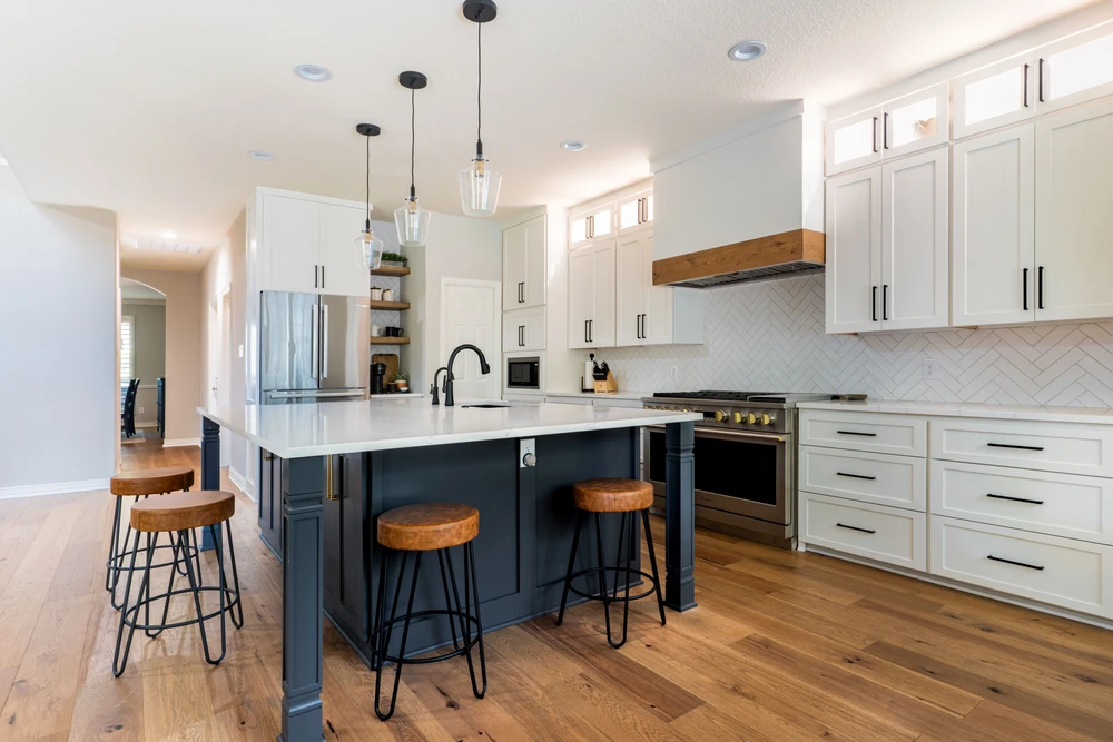 A spacious modern kitchen with a dark blue island and bar stools, white cabinetry, stainless steel appliances, and wood floors.