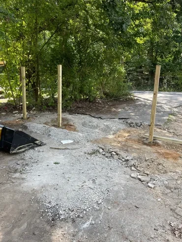 Four wooden posts stand amidst gravel, dirt, and broken pavement, backed by lush trees, indicating a new fence or gate project.