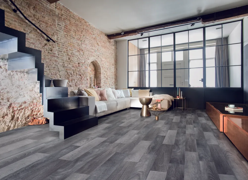 Modern living room with a large L-shaped sofa, exposed brick wall, industrial windows, and dark grey wood flooring.