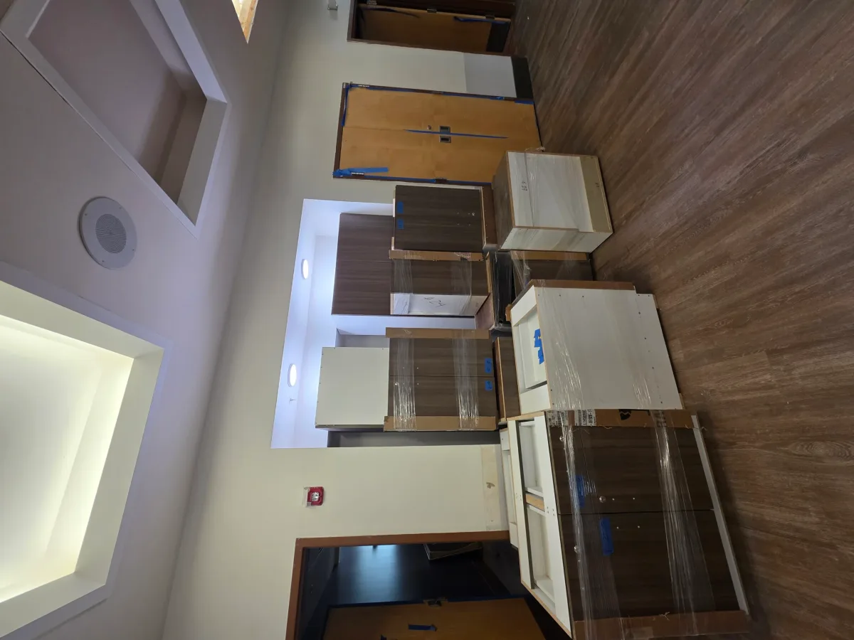 Modern room with wood flooring, light walls, recessed lighting, and numerous stacked, plastic-wrapped white and brown cabinet units.