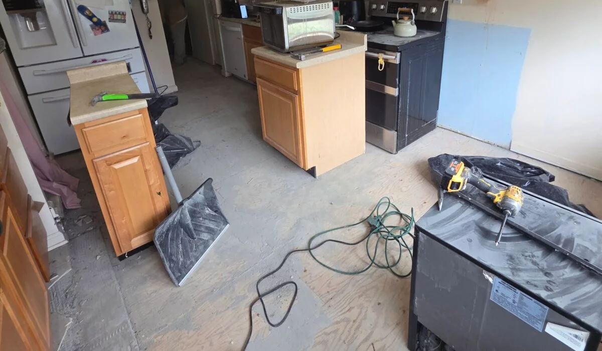 A kitchen mid-renovation featuring appliances, displaced cabinets, a hammer, drill, and exposed subfloor.