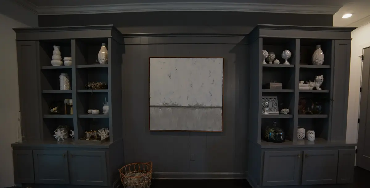 Two dark gray built-in bookshelves filled with decorative objects flank an abstract white and gray painting on a gray wall.