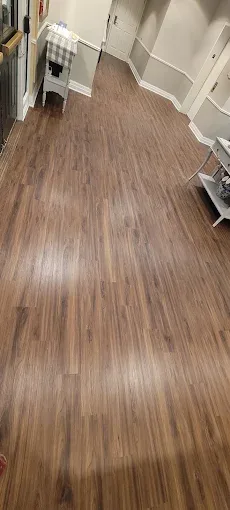 A spacious hallway features long planks of warm brown wood-look laminate flooring, with light walls and white trim.