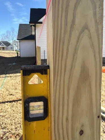 A yellow spirit level checks the vertical alignment of a wooden post, with a red string line and new house construction visible.