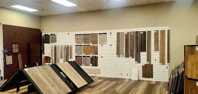 Interior view of a bright flooring showroom, featuring various wood-look plank and square tile flooring samples on wall displays and floor racks.