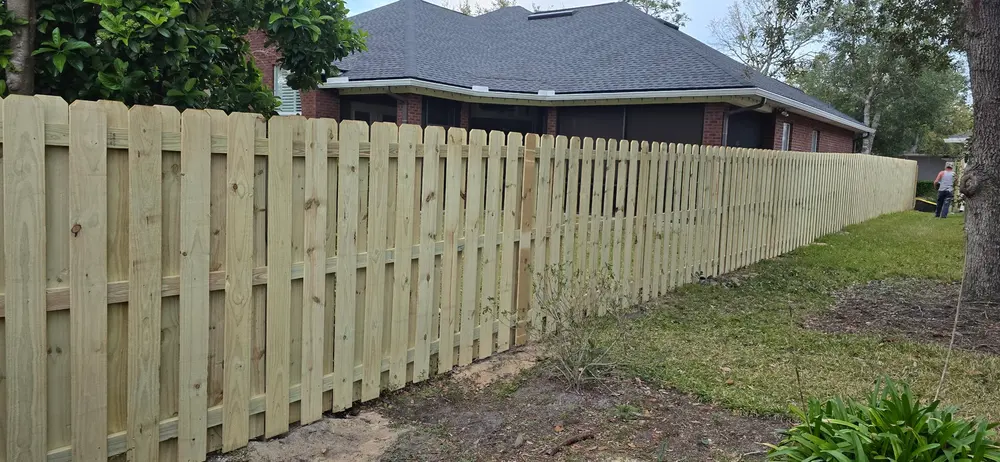 A newly installed light-colored wooden privacy fence with dog-eared pickets stretches across a green yard towards a house.
