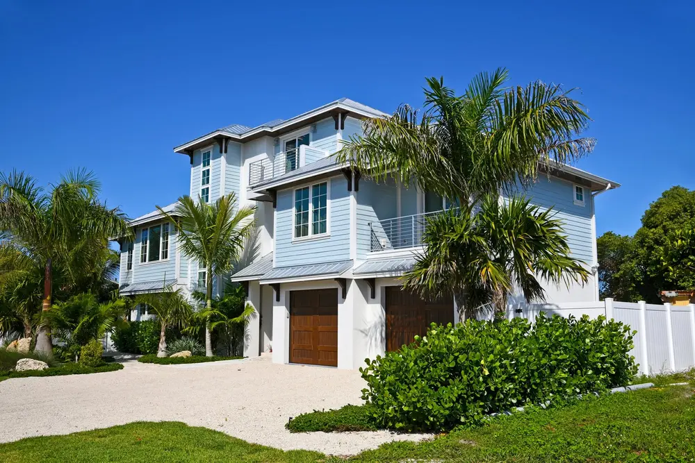 Modern light blue multi-story coastal house with balconies, dark garage doors, tropical palm trees, and a gravel driveway.