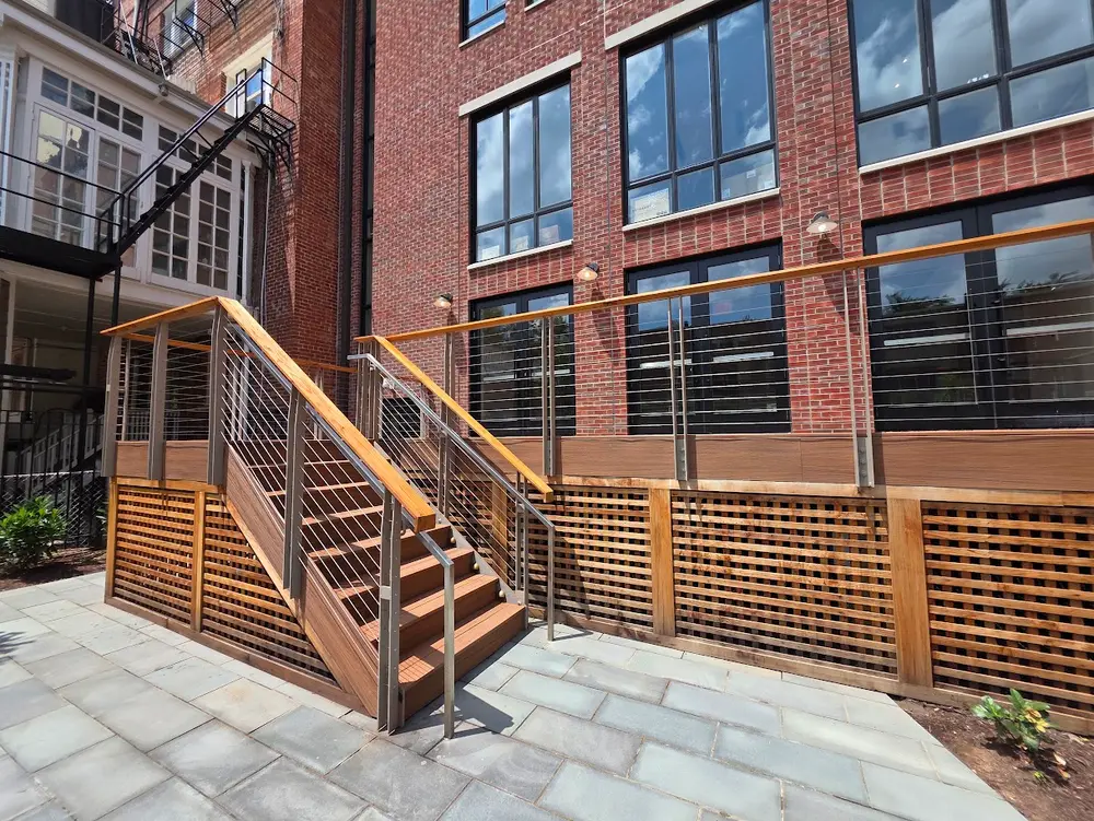 Modern wood and steel cable-railing stairs ascend to a lattice-skirted deck beside a brick building and flagstone patio.