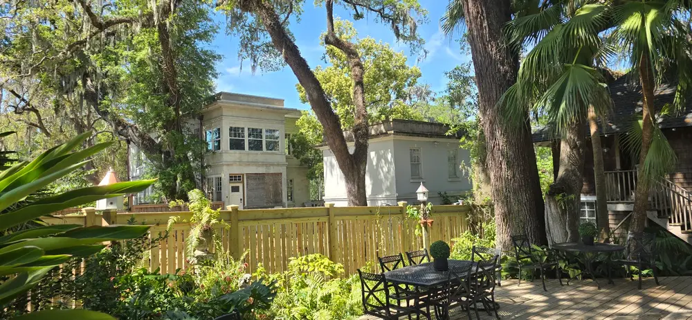 Lush backyard with an outdoor dining set, wooden fence, Spanish moss trees, and a modern house in the background.