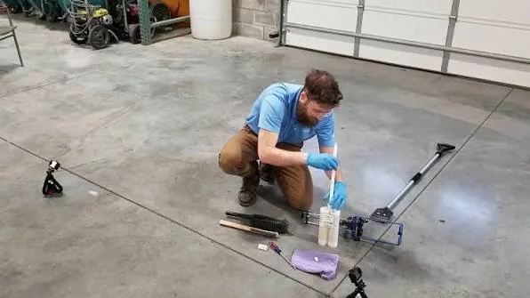 Bearded man in blue shirt and gloves kneels on concrete floor, mixing materials for a project.