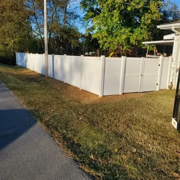 A long, new white vinyl privacy fence with two gates encloses a residential backyard alongside a paved path on a sunny day.