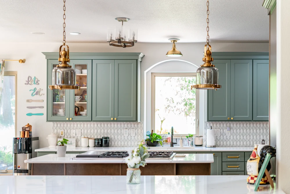 A bright, modern kitchen features sage green cabinets, brass hardware, a white geometric backsplash, a kitchen island, and stylish pendant lights.