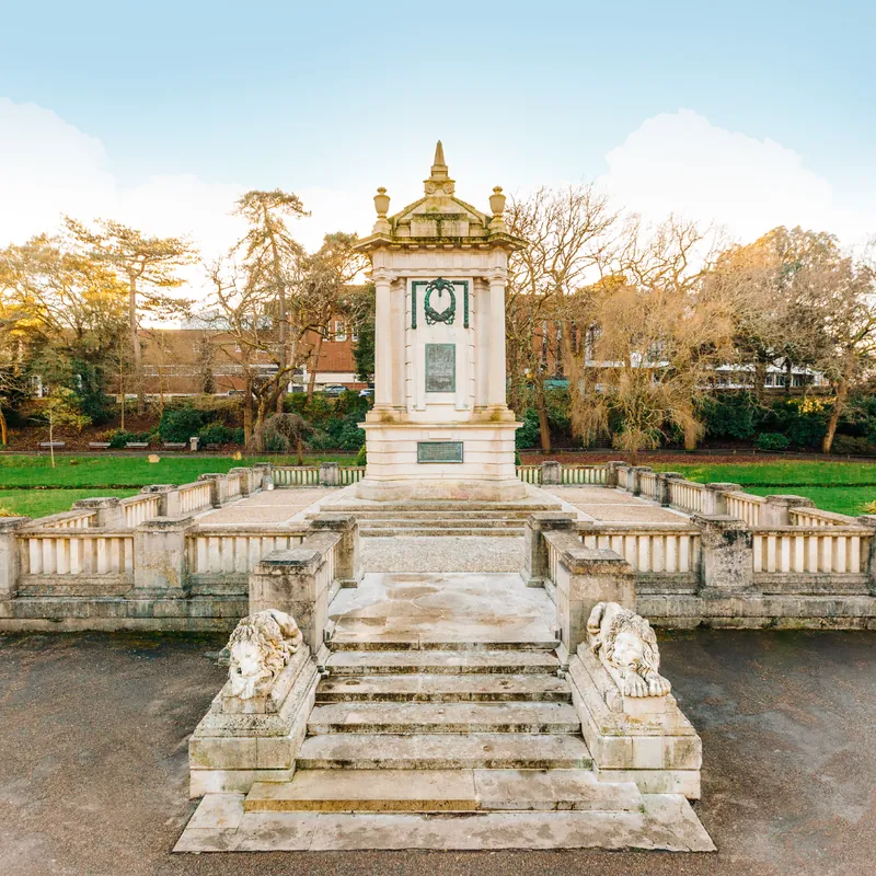 Tall, ornate golden monument structure standing in a park setting against a pale blue sky
