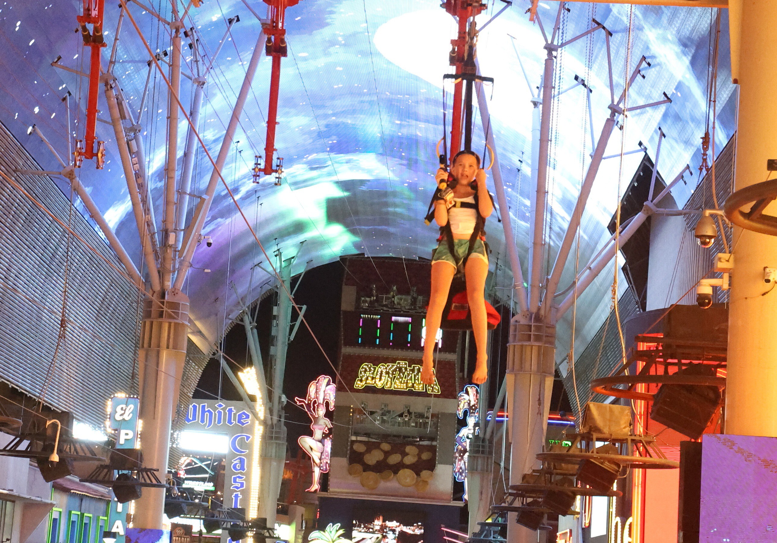 A girl rides SlotZilla seated in a zipline harness, legs dangling, under the glowing Fremont Street dome in Las Vegas