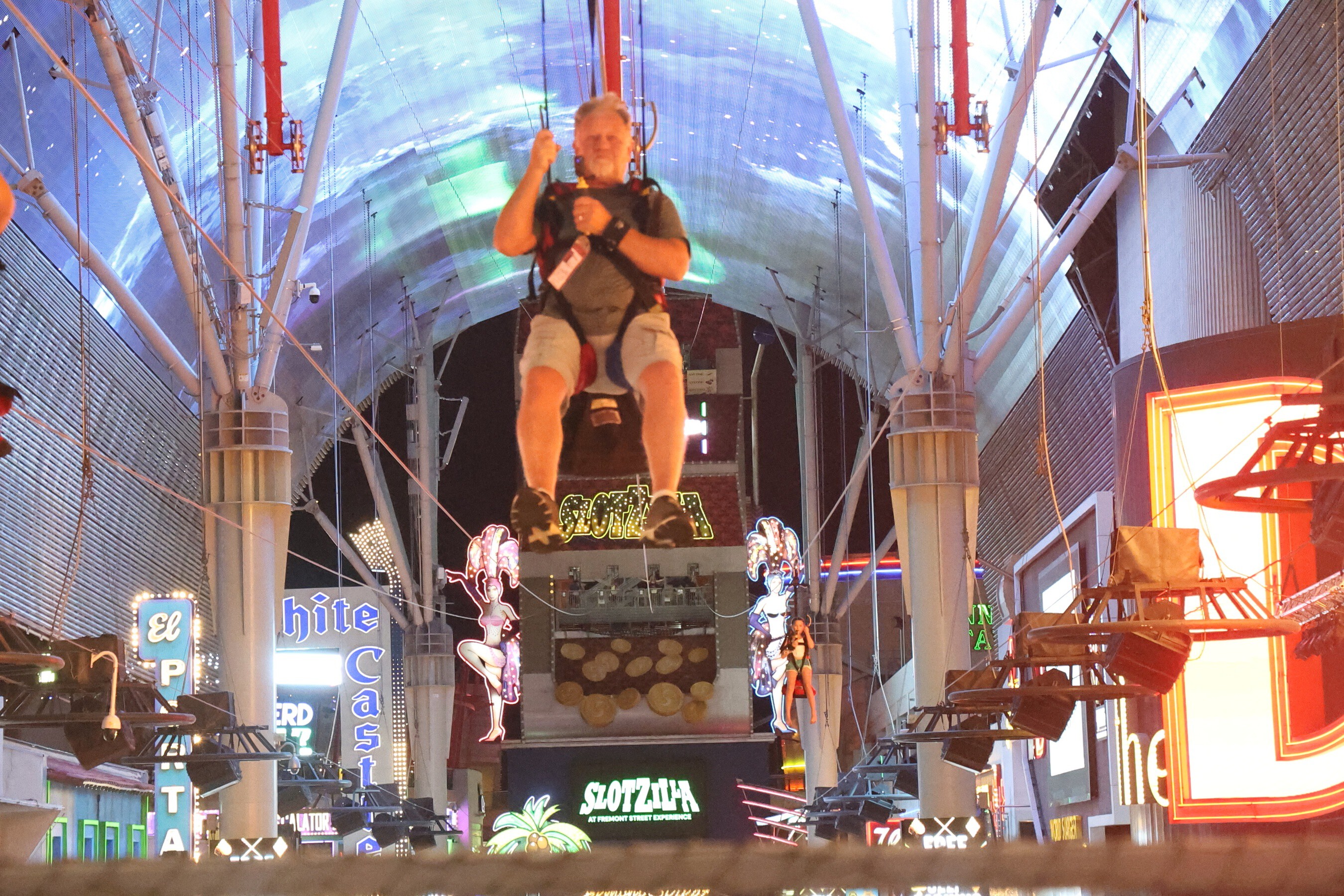 A man rides SlotZilla seated in a zipline harness, legs dangling, under the glowing Fremont Street dome in Las Vegas