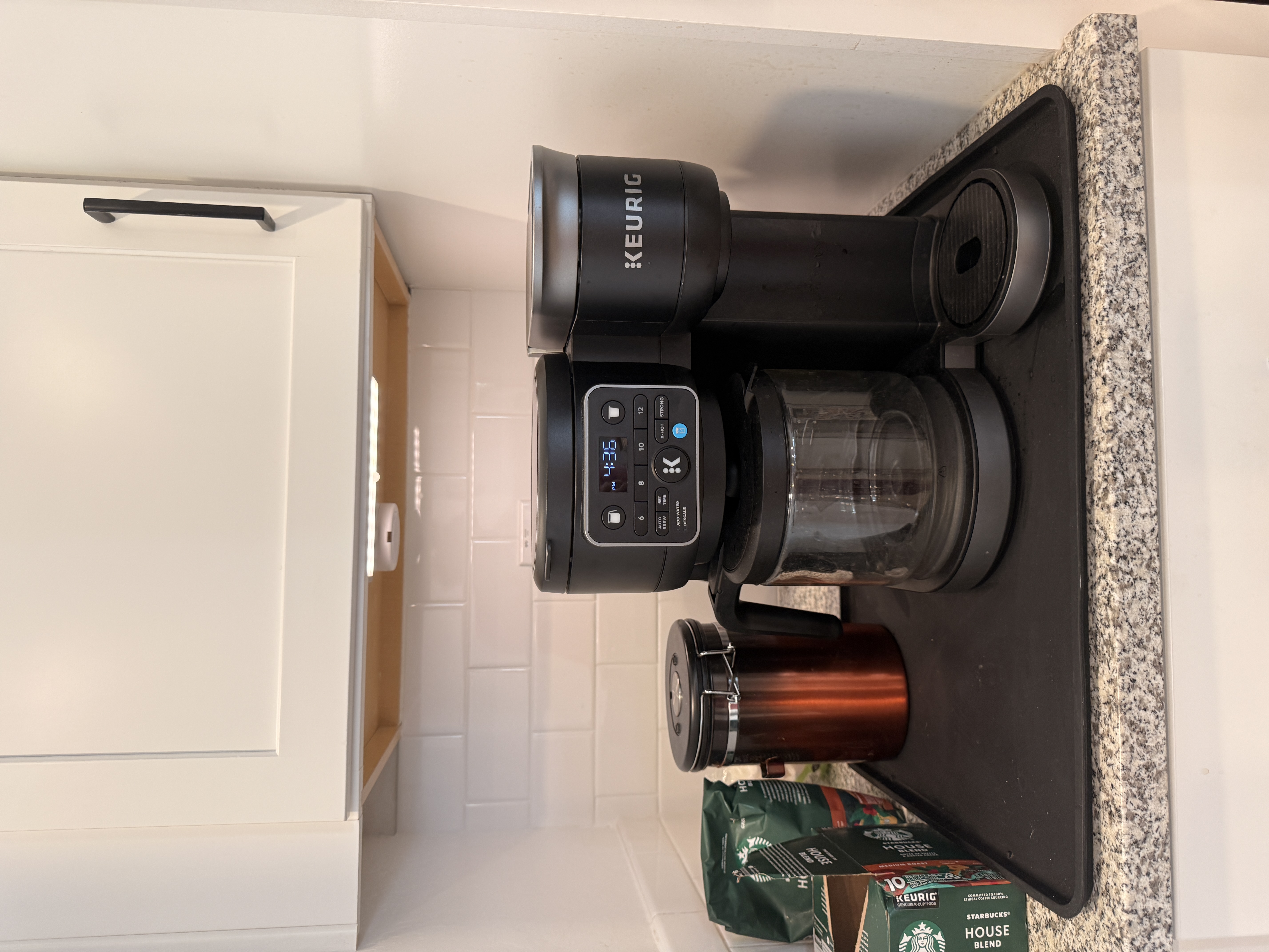 Keurig coffee maker and supplies on a black countertop tray beneath an illuminated cabinet. Keurig coffee maker and supplies on a black countertop tray beneath an illuminated cabinet.