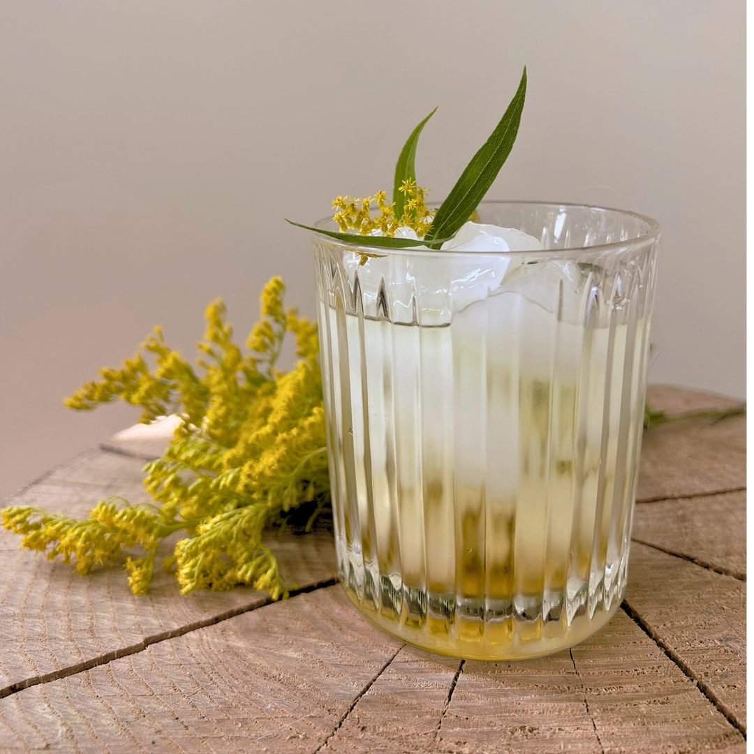 Goldenrod simple syrup cocktail with ice and fresh goldenrod garnish served in a glass on a wooden surface.
