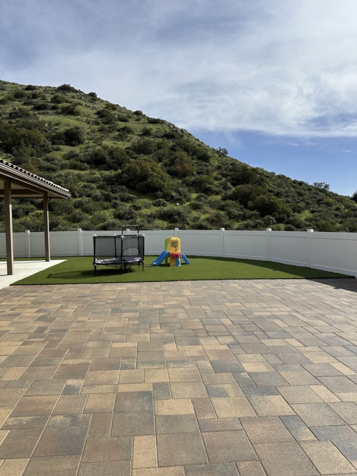 Paver patio with turf play area, trampoline, and slide set against a hillside