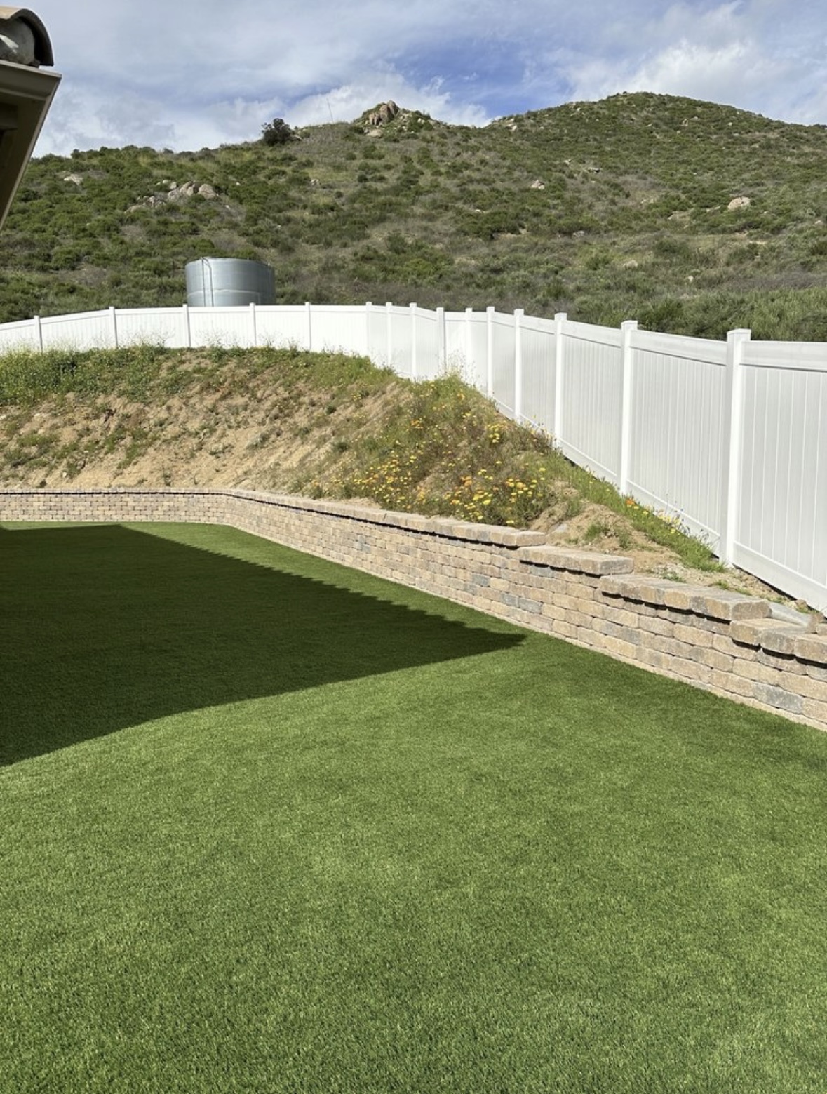 Curved stone retaining wall holding back a hillside, with new artificial turf and white vinyl fencing