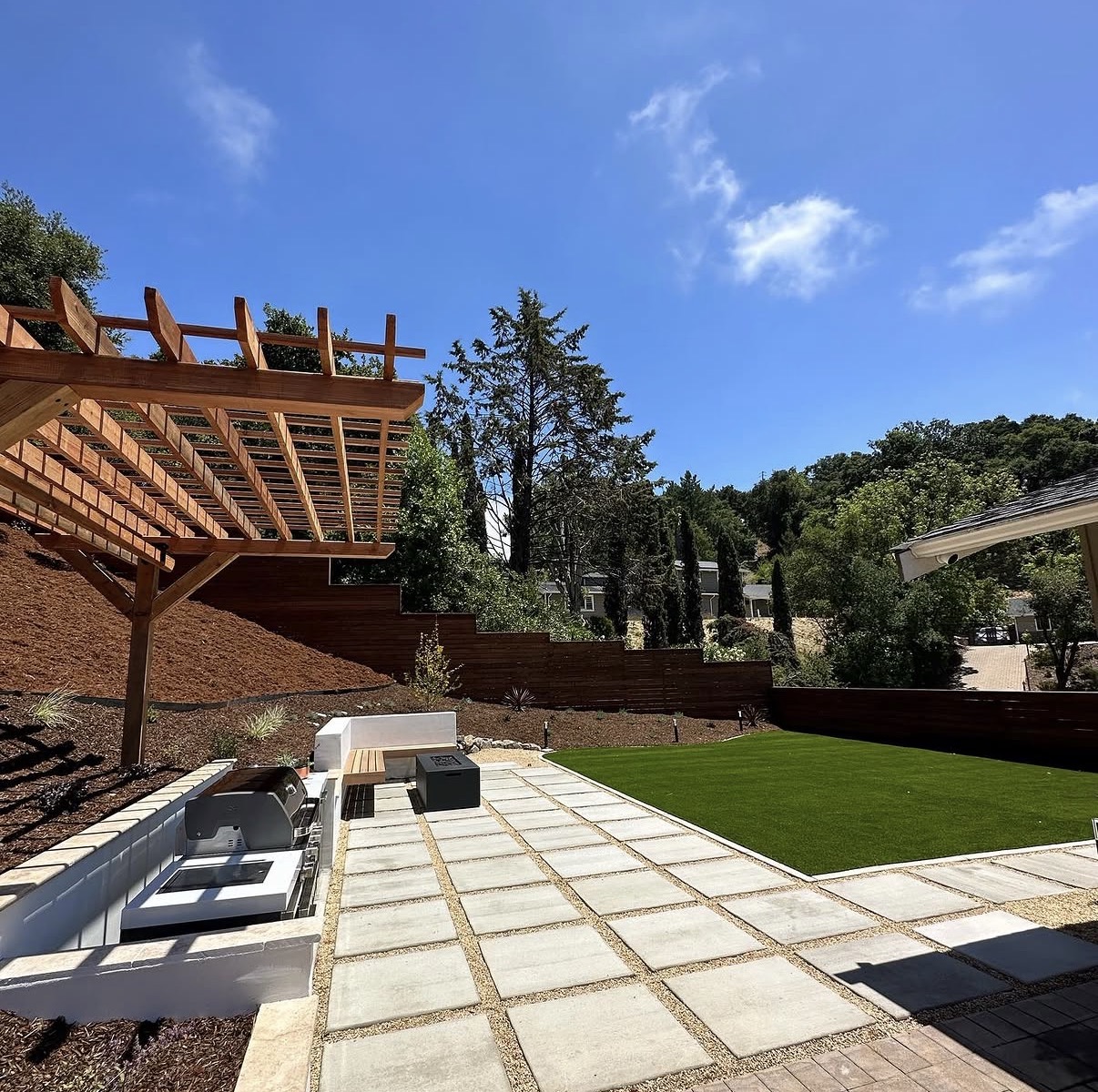 Backyard outdoor kitchen with pergola, large format pavers, artificial turf, and wood privacy fence