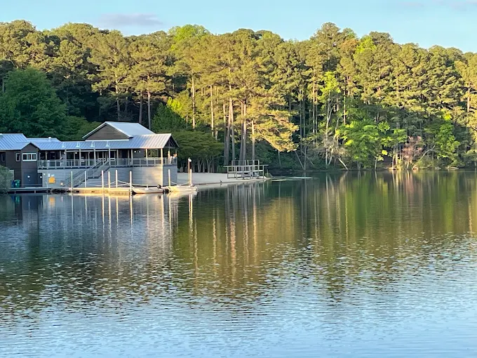 A serene lakeside view at Lake Johnson Park with a marina building, dock, and sandy beach, surrounded by dense trees. 