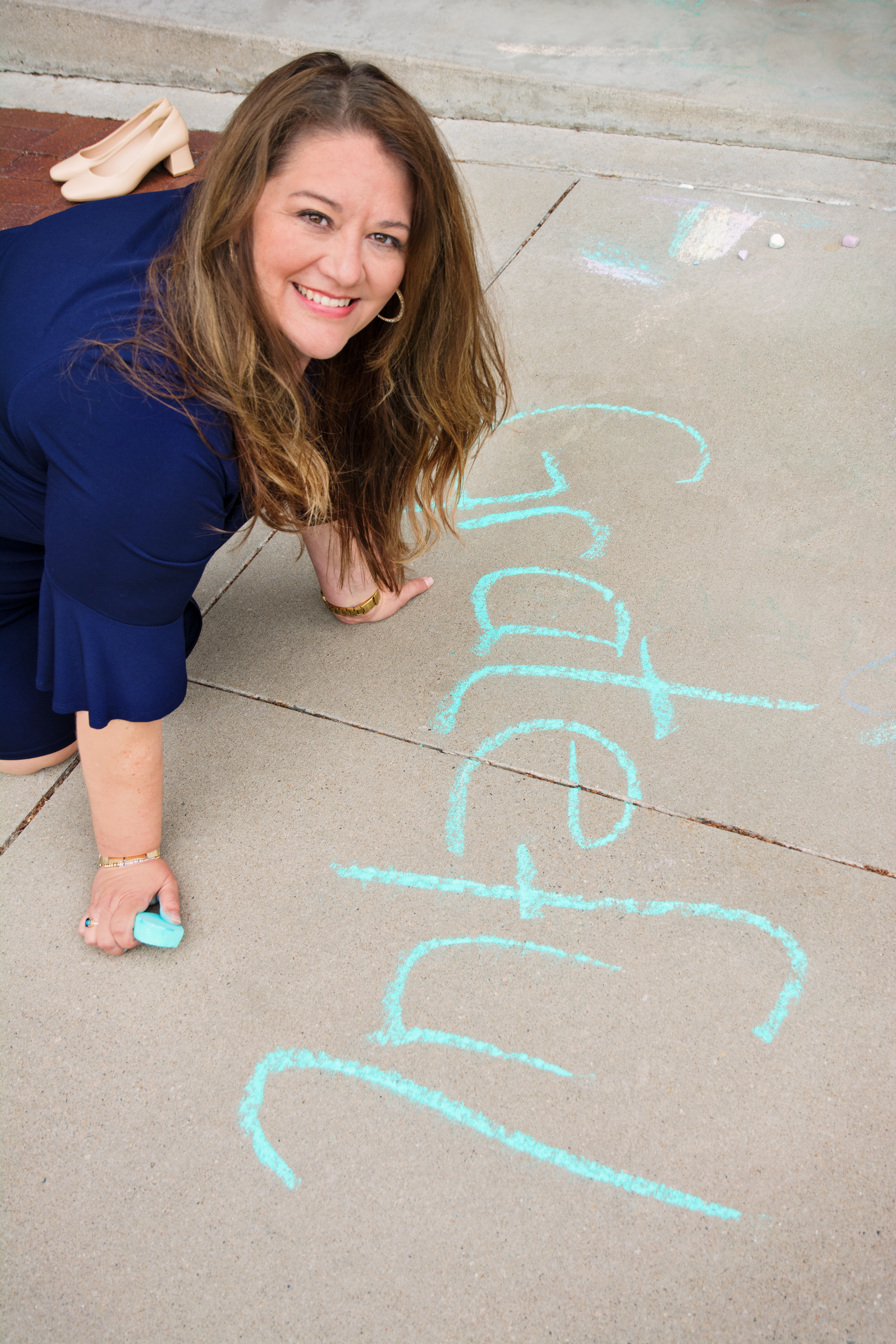 Wendy Hoerl kneeling on a sidewalk, smiling, writing the word Grateful in chalk