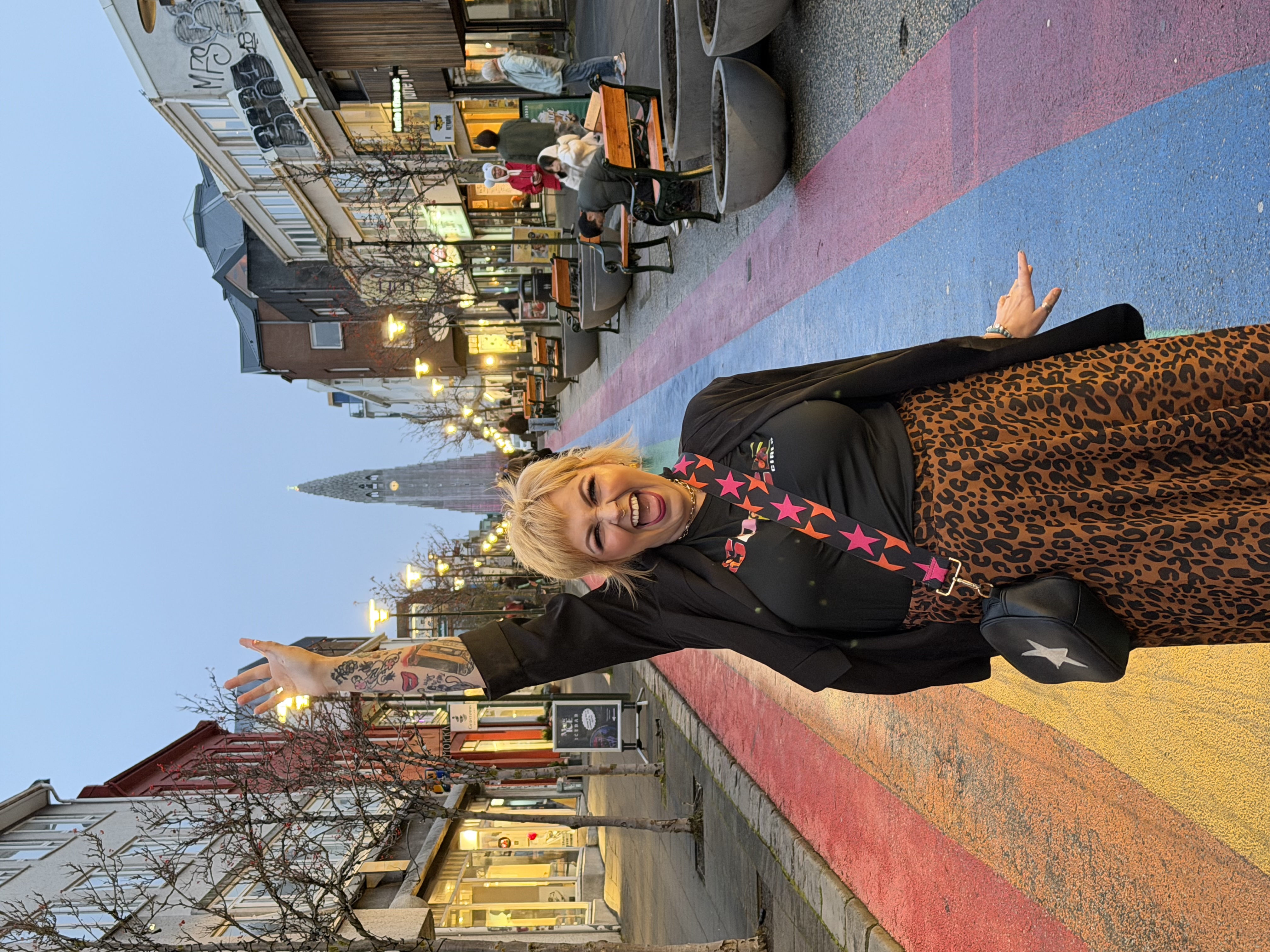 Kim Antrobus laughing with one arm in the air on a rainbow painted street
