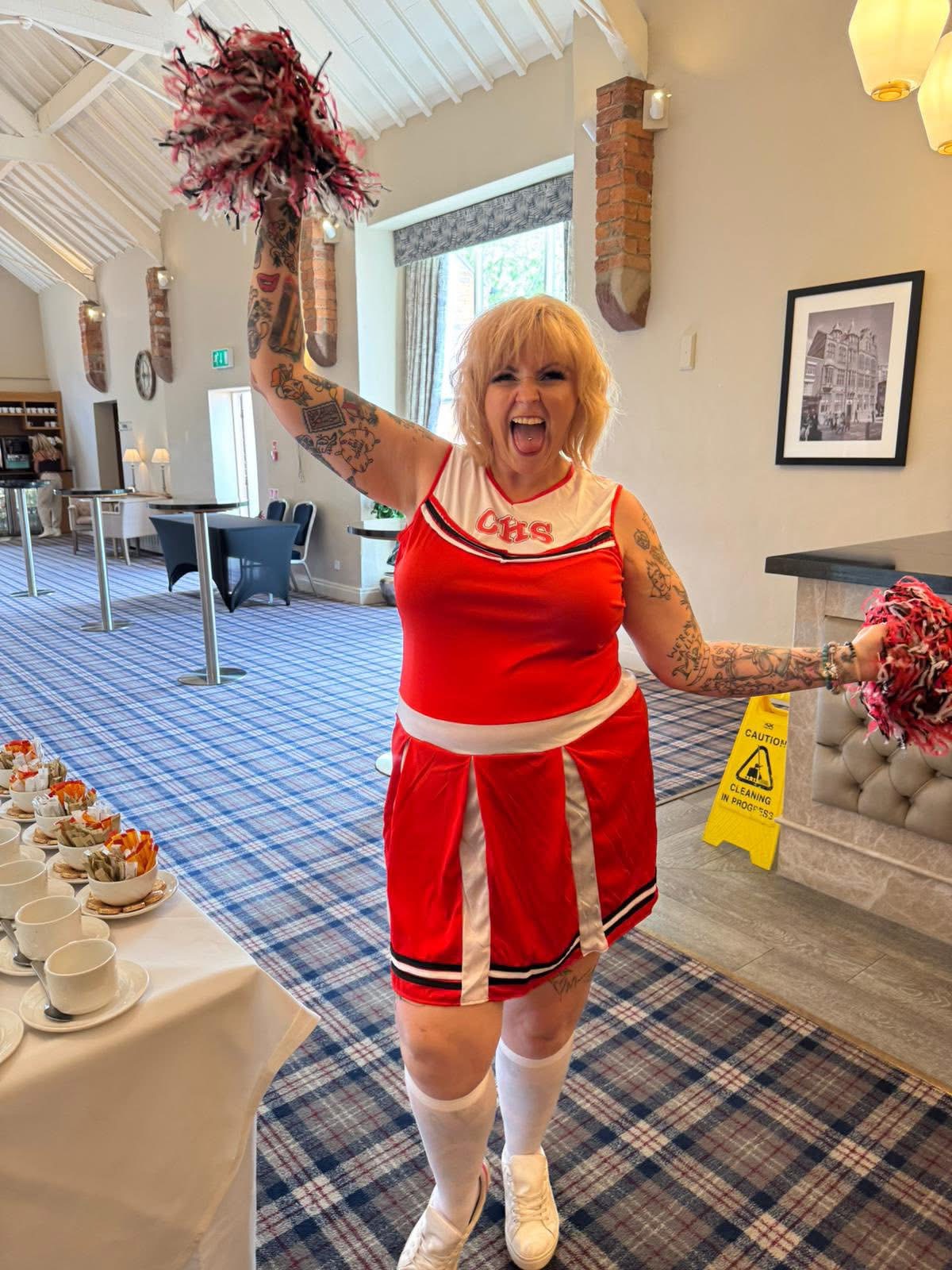 Kim Antrobus in a red cheerleader outfit, shouting with joy and waving pom poms