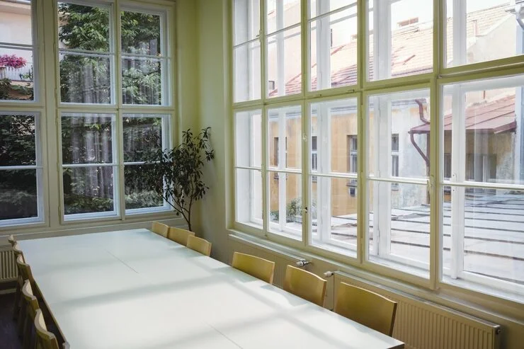 Une longue table blanche vide avec des chaises dans une pièce baignée de lumière, dotée de grandes fenêtres donnant sur une cour intérieure d’immeuble.