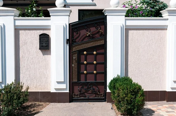 Un portail en métal orné, brun foncé et noir, partiellement ouvert, avec des accents en bronze, encadré par des piliers blancs et un mur rose pâle.