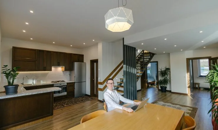 Une femme travaille sur un ordinateur portable à une table en bois clair dans une maison moderne avec une cuisine ouverte et un élégant escalier design.