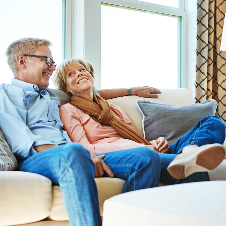 Un couple de seniors heureux se détend sur un canapé beige, souriant et regardant vers le haut, baigné par la lumière naturelle entrant par la fenêtre.