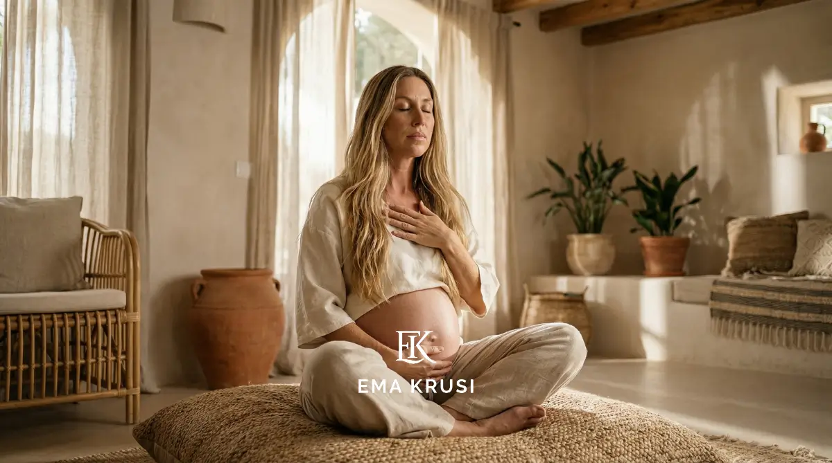 Femme enceinte assise en tailleur, main sur le cœur et sur le ventre, en méditation dans un intérieur aux tons naturels.