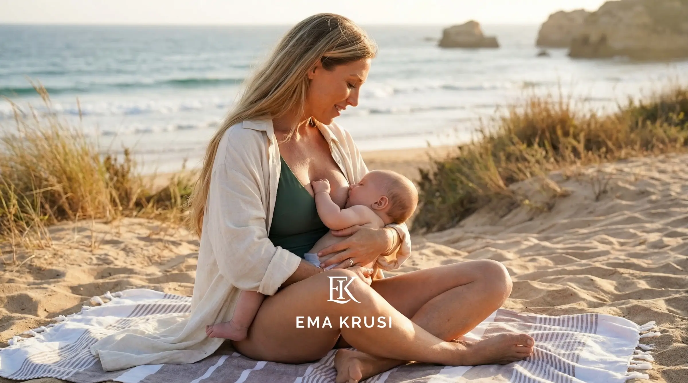 Femme allaitant son bébé sur une plage au coucher du soleil, assise sur une serviette face à l’océan.