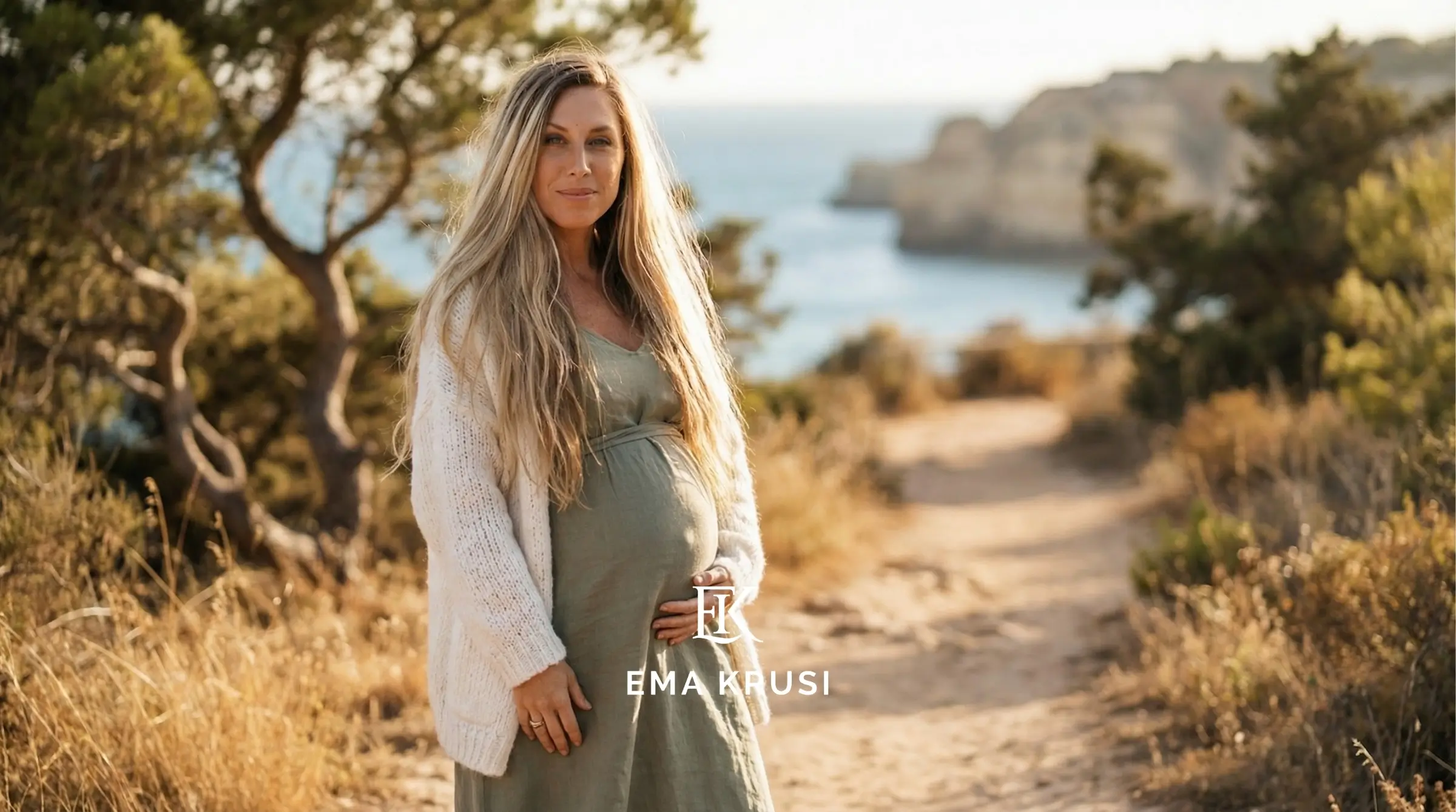 Femme enceinte debout sur un sentier en bord de mer, main sur le ventre, lumière de fin de journée.