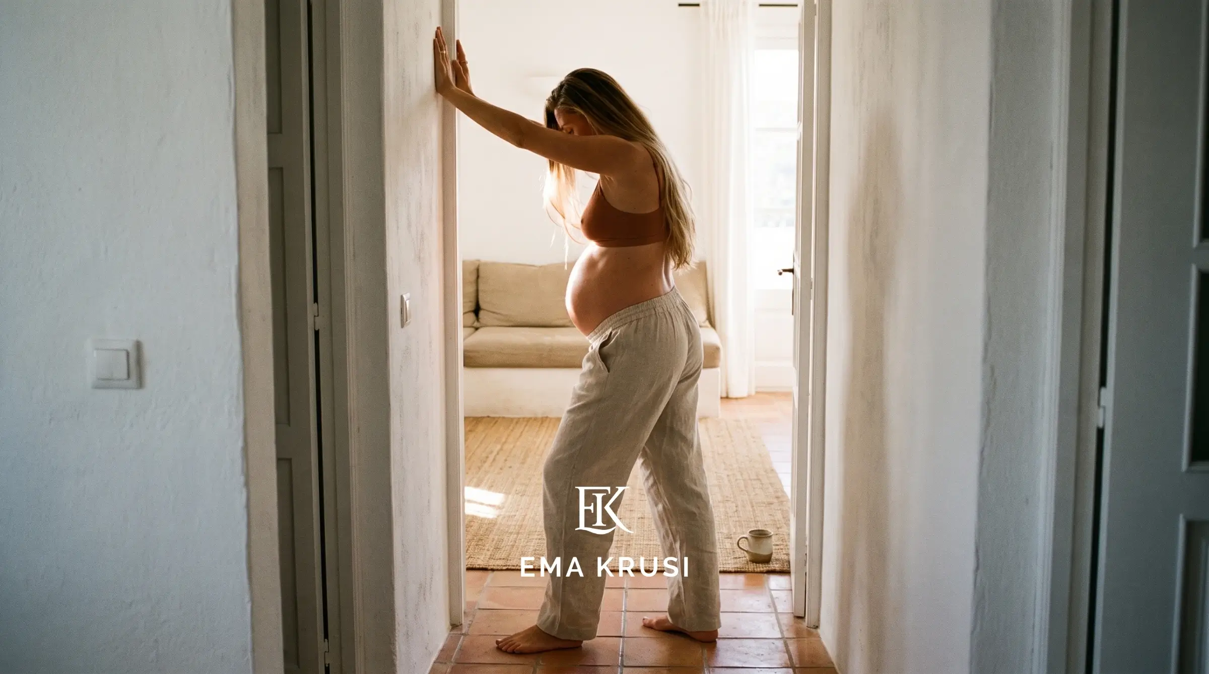 Une femme enceinte debout dans l'encadrement d'une porte, une main appuyée au mur, la tête baissée, chez elle en pleine lumière du jour.