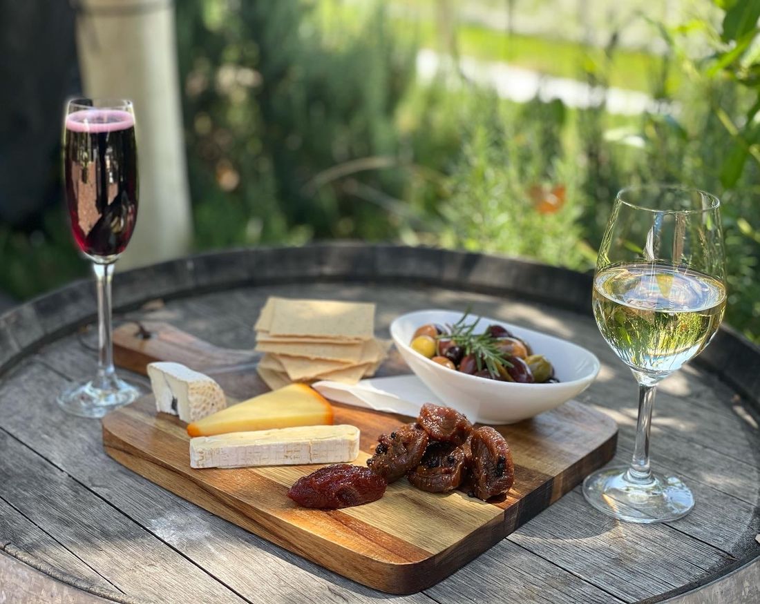 A cheese board with olives and quince paste on a wine barrel table, with two glasses of wine and the garden behind