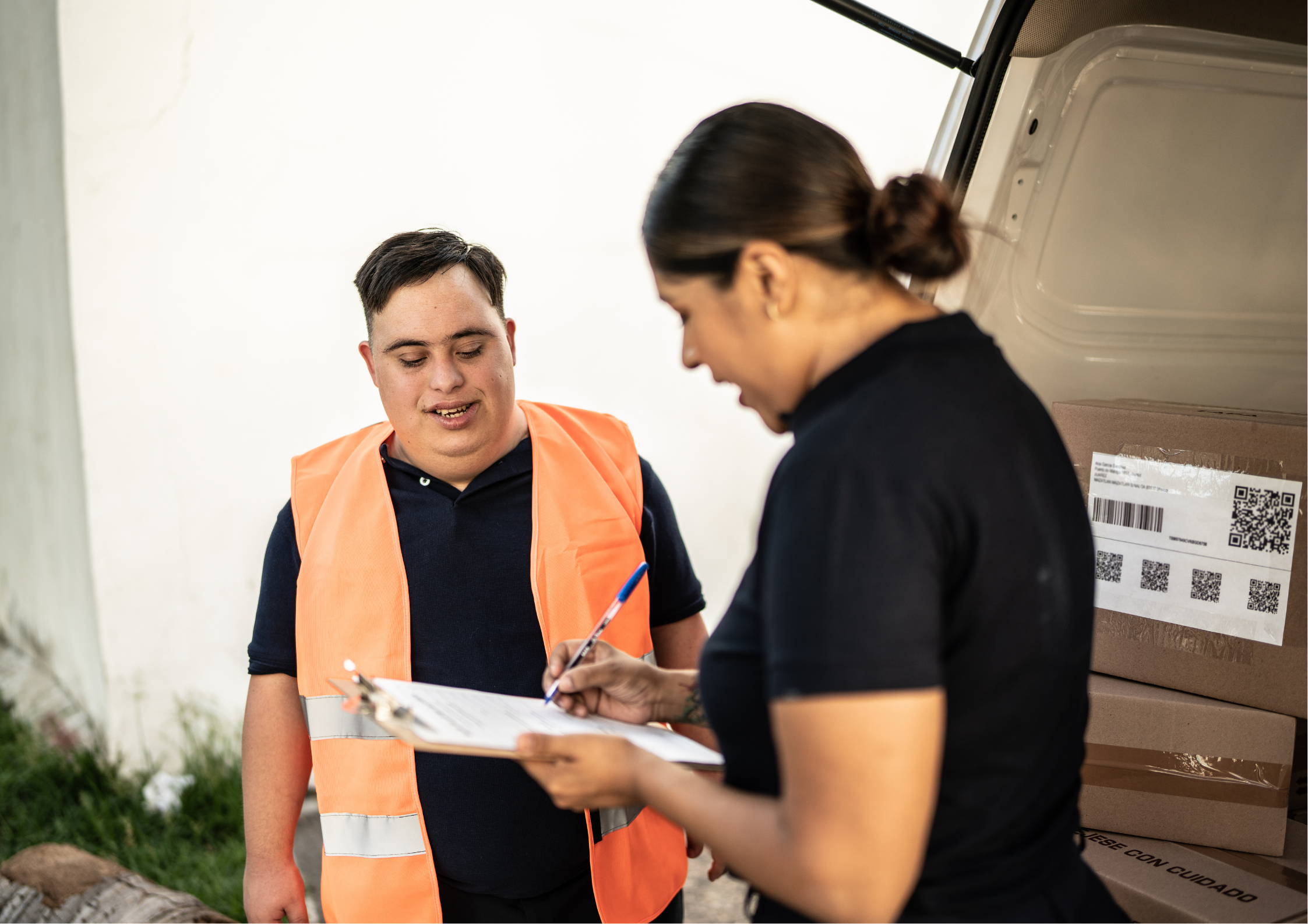 A young man with Down syndrome wearing an orange safety vest assists a coworker signing a clipboard next to a delivery van filled with packages.