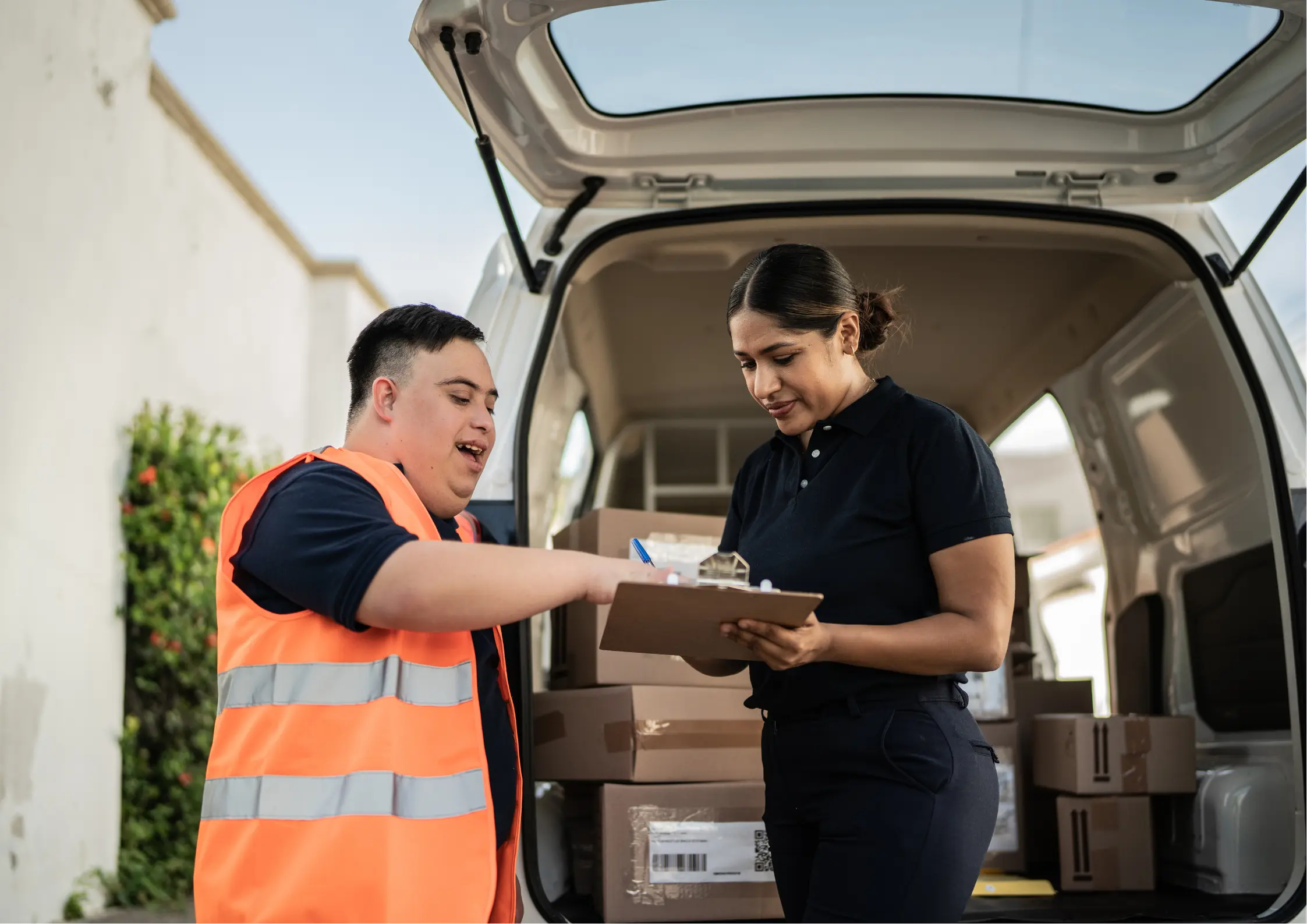 A young adult worker with Down syndrome wearing a safety vest filling out paperwork on a clipboard with a coworker near a delivery van. A young adult worker with Down syndrome wearing a safety vest filling out paperwork on a clipboard with a coworker near a delivery van.