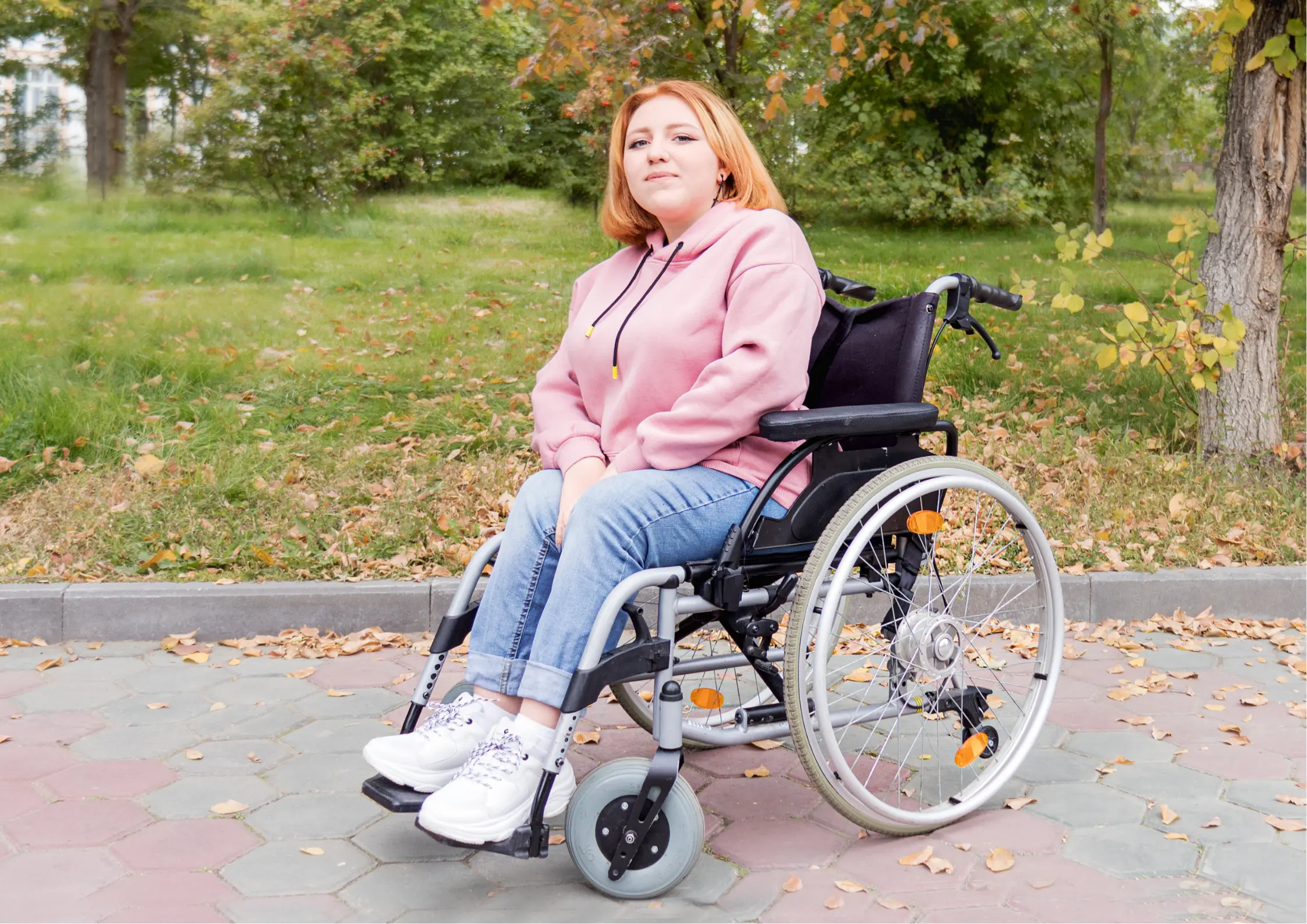 Woman in a pink hoodie sitting in her wheelchair on a park path surrounded by autumn leaves