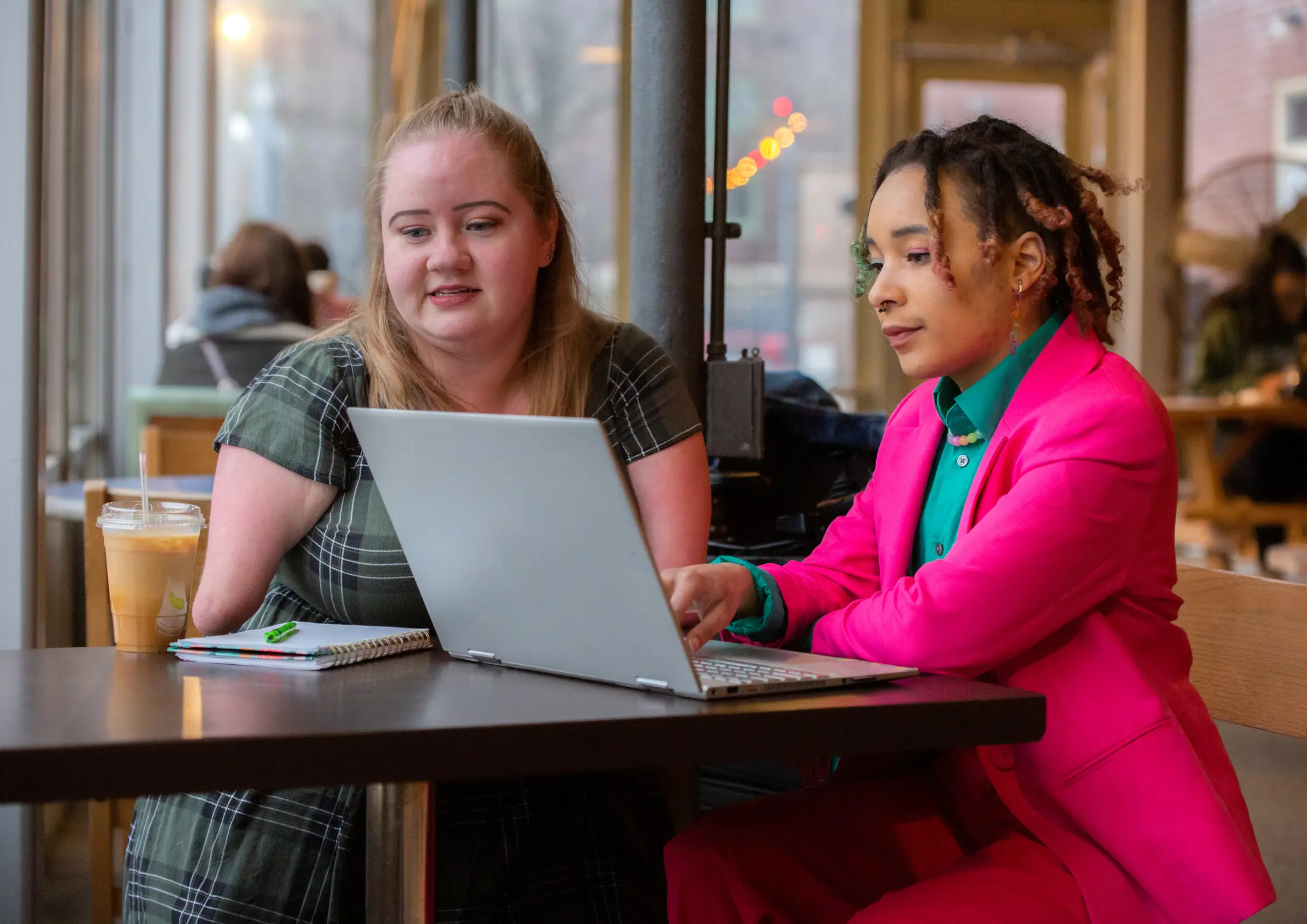 Two women at a café table working together on a laptop with a notebook nearby Two women at a café table working together on a laptop with a notebook nearby