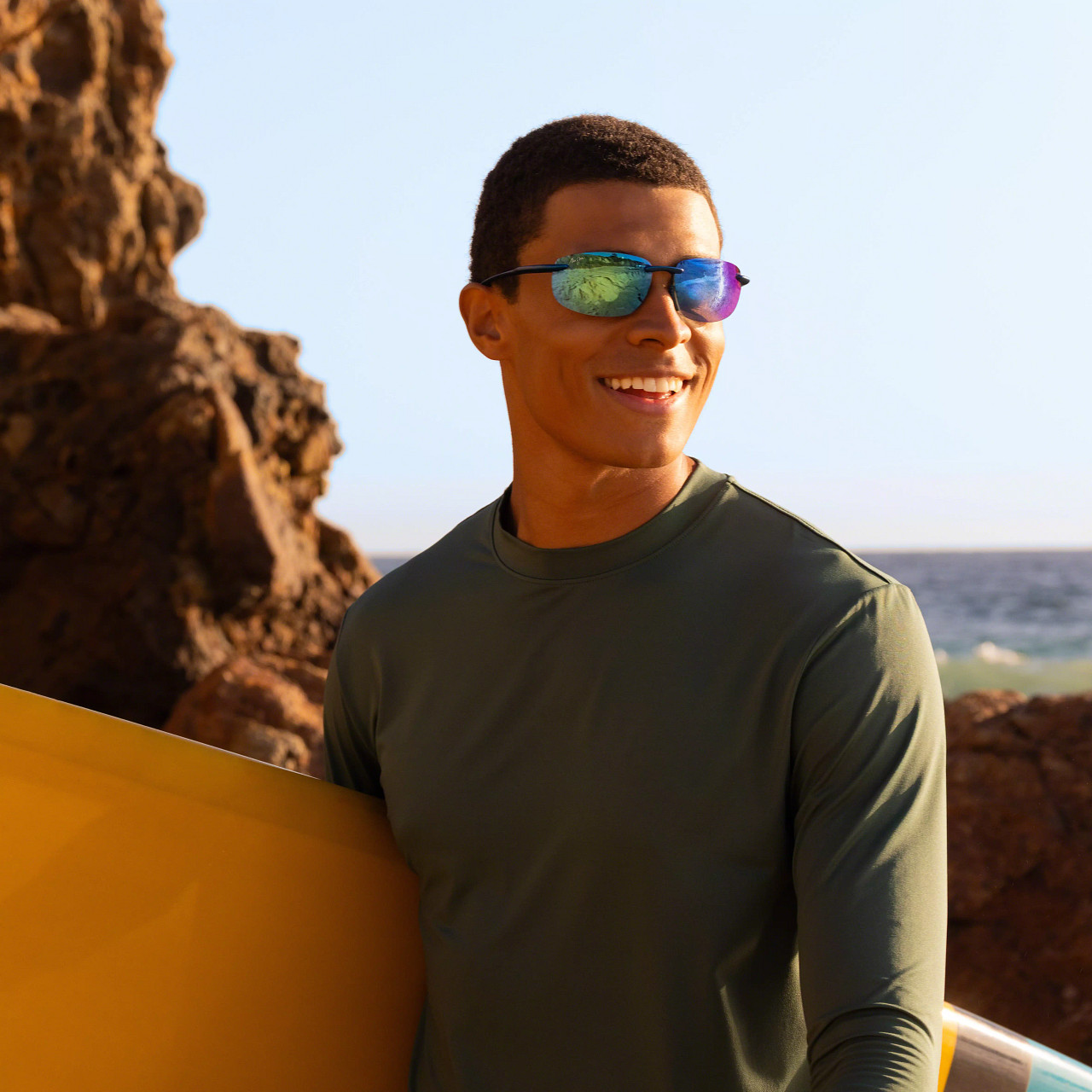 Man wearing mirrored Maui Jim sunglasses and holding a surfboard beside the ocean. Man wearing mirrored Maui Jim sunglasses and holding a surfboard beside the ocean.