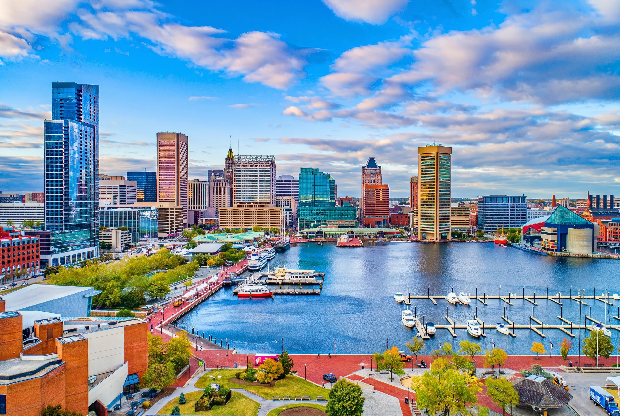 Baltimore's Inner Harbor with downtown towers, docked boats and the waterfront promenade under a partly cloudy sky